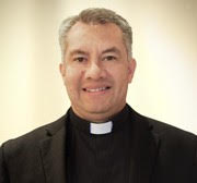 A priest in a black suit smiles for the camera.