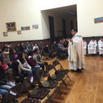 A priest speaks to a group of people in a room.