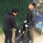 Two men standing next to a trash bag on a sidewalk.