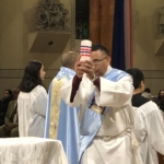 A priest holds up a cup in front of a group of people.