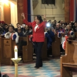 A woman in a red jacket is holding a flag in front of a church.