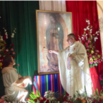 A priest is standing in front of a painting of guadalupe.