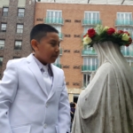 A boy in a white suit standing next to a statue of the virgin mary.