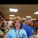 A woman in a blue shirt standing in a room full of people.