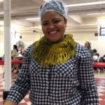 A woman wearing a yellow hat and a yellow scarf standing in a kitchen.